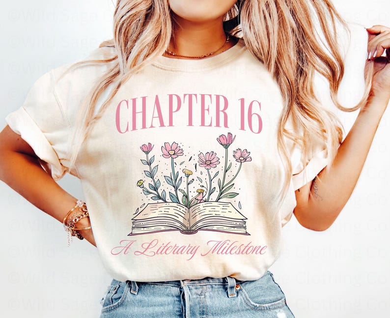 Puede incluir: Una camiseta de color crema con el texto "CHAPTER 16 A Literary Milestone" en rosa. La camiseta presenta un dise&ntilde;o floral rosa con un libro en el centro.
