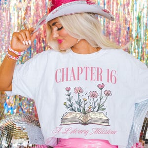 Puede incluir: Camiseta blanca con texto rosa que dice "Cap&iacute;tulo 16 Un hito literario". La camiseta presenta un dibujo de un libro abierto con flores rosas.
