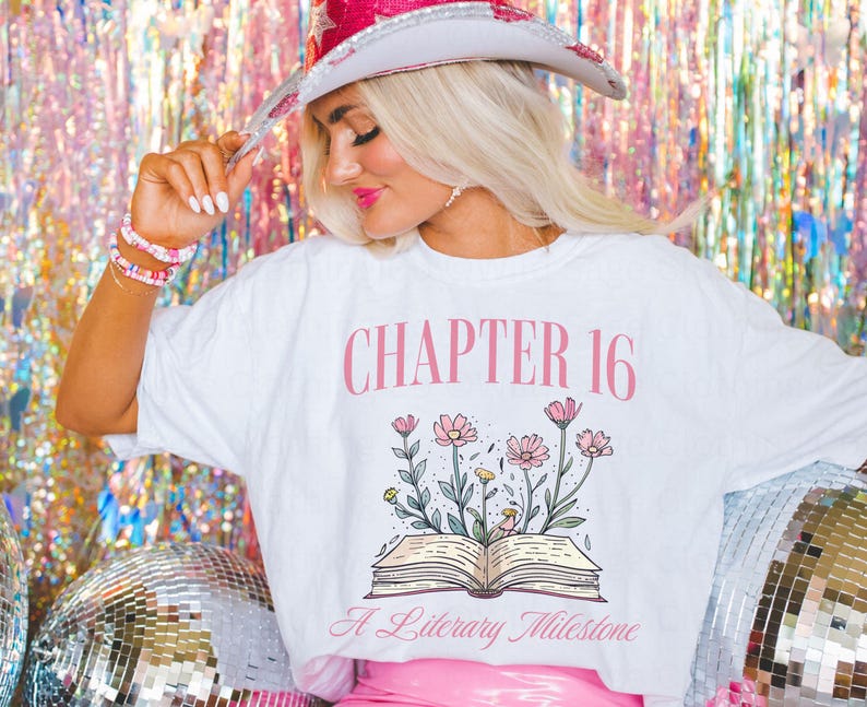 Puede incluir: Camiseta blanca con texto rosa que dice "Cap&iacute;tulo 16 Un hito literario". La camiseta presenta un dibujo de un libro abierto con flores rosas.