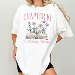 Puede incluir: Camiseta blanca de gran tama&ntilde;o con un gr&aacute;fico rosa de un libro abierto con flores y el texto "Cap&iacute;tulo 16 Un hito literario".
