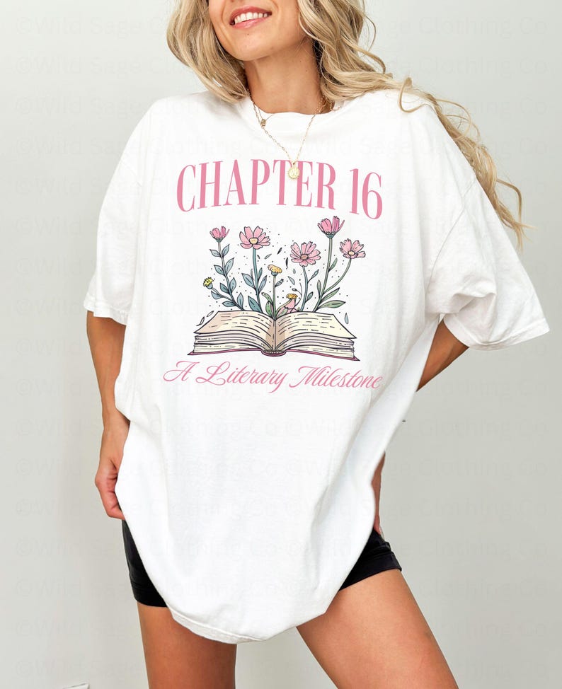 Puede incluir: Camiseta blanca de gran tama&ntilde;o con un gr&aacute;fico rosa de un libro abierto con flores y el texto "Cap&iacute;tulo 16 Un hito literario".