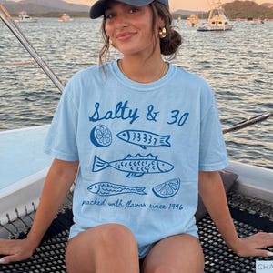 May include: Pale blue t-shirt featuring the words "Salty & 30" and fish and lemon illustrations. The shirt also has the text "packed with flavour since 1996".