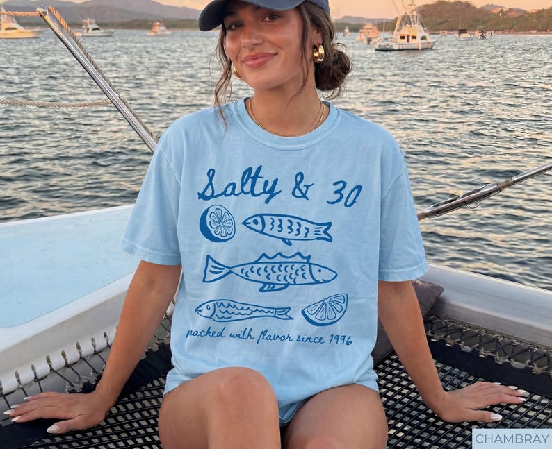 May include: Pale blue t-shirt featuring the words "Salty & 30" and fish and lemon illustrations. The shirt also has the text "packed with flavour since 1996".