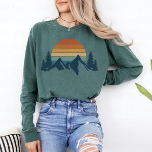 May include: A teal long-sleeved shirt with a vintage-style sunset graphic featuring mountains and trees. The graphic is in shades of orange, yellow, and blue.