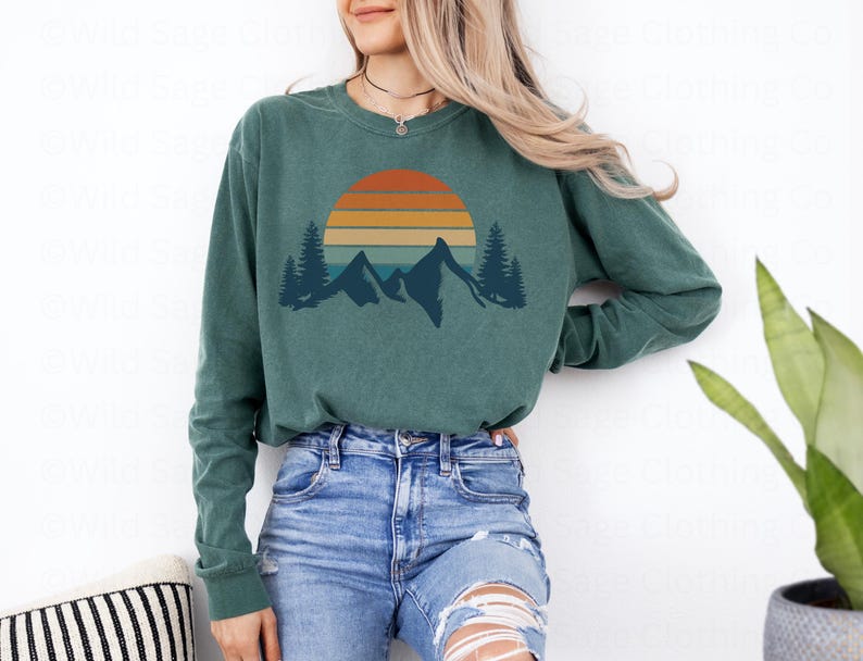 May include: A teal long-sleeved shirt with a vintage-style sunset graphic featuring mountains and trees. The graphic is in shades of orange, yellow, and blue.