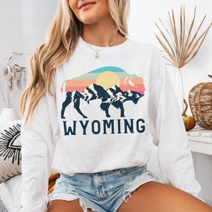 May include: White long-sleeve sweatshirt with a graphic of a bison silhouette in front of a mountain range and a sunset. The text "WYOMING" is printed below the graphic.