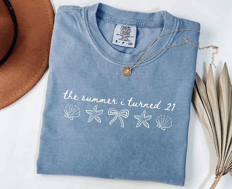 Pu&ograve; includere: Una maglietta blu Comfort Colors con la scritta "the summer i turned 21" e grafiche di conchiglie, stelle marine e fiocchi. Una collana d'oro con un ciondolo &egrave; drappeggiata sulla maglietta.