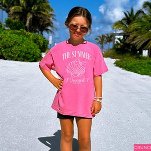 K&ouml;nnte beinhalten: Ein leuchtend pinkfarbenes T-Shirt mit dem Text "THE SUMMER I Turned 7" und einer Muschel-Grafik. Das T-Shirt wird von einer Person getragen, die eine Sonnenbrille und Armb&auml;nder tr&auml;gt und auf einem sandigen Weg mit Palmen im Hintergrund steht.