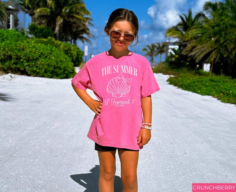 K&ouml;nnte beinhalten: Ein leuchtend pinkfarbenes T-Shirt mit dem Text "THE SUMMER I Turned 7" und einer Muschel-Grafik. Das T-Shirt wird von einer Person getragen, die eine Sonnenbrille und Armb&auml;nder tr&auml;gt und auf einem sandigen Weg mit Palmen im Hintergrund steht.