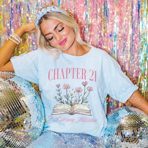 May include: A light blue t-shirt with the text "CHAPTER 21 A Literary Milestone" and a floral design with a book underneath. The shirt is worn by a person with a headband and is posed against a sparkly background.
