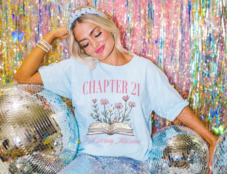 May include: A light blue t-shirt with the text "CHAPTER 21 A Literary Milestone" and a floral design with a book underneath. The shirt is worn by a person with a headband and is posed against a sparkly background.