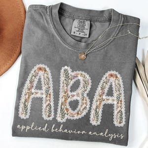 May include: A gray Comfort Colors t-shirt with the letters "ABA" in a floral design, outlined with white lace. The words "applied behavior analysis" are printed below the letters. A gold necklace rests on the shirt.