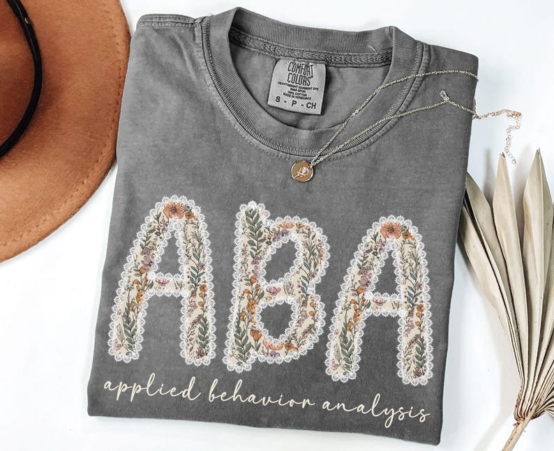 May include: A gray Comfort Colors t-shirt with the letters "ABA" in a floral design, outlined with white lace. The words "applied behavior analysis" are printed below the letters. A gold necklace rests on the shirt.