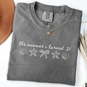 Pu&ograve; includere: Una maglietta grigia Comfort Colors con la scritta "the summer i turned 21" e grafiche di conchiglie, stelle marine e fiocchi. Una collana d'oro e un cappello marrone sono anche nell'immagine.