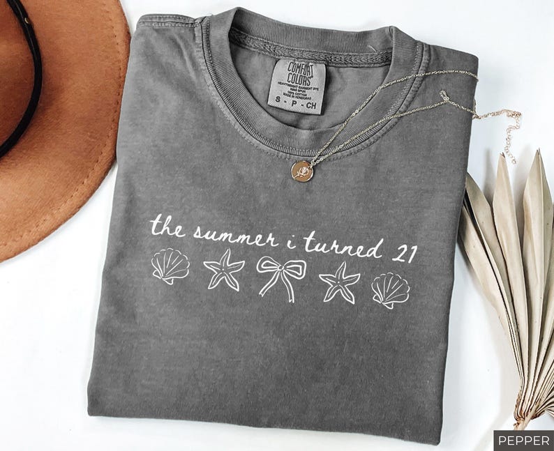 Pu&ograve; includere: Una maglietta grigia Comfort Colors con la scritta "the summer i turned 21" e grafiche di conchiglie, stelle marine e fiocchi. Una collana d'oro e un cappello marrone sono anche nell'immagine.