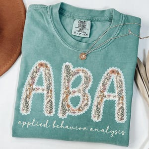 May include: A teal Comfort Colors t-shirt with the letters "ABA" in a floral design. The words "applied behavior analysis" are written below the letters in a script font. A silver necklace with a pendant is on the shirt.