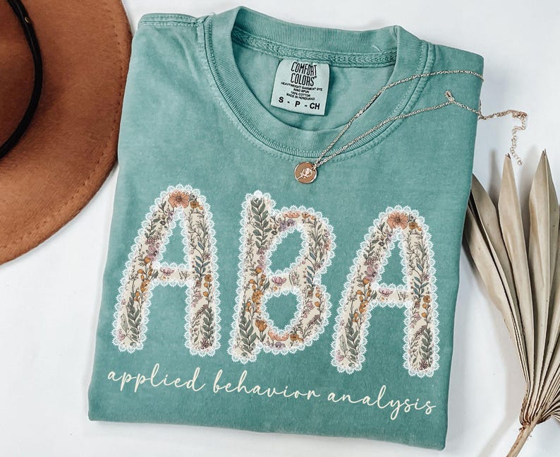 May include: A teal Comfort Colors t-shirt with the letters "ABA" in a floral design. The words "applied behavior analysis" are written below the letters in a script font. A silver necklace with a pendant is on the shirt.
