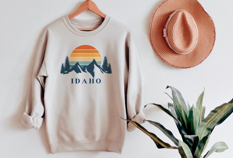 May include: A beige sweatshirt with a graphic print of mountains, a sunset, and trees. The text "IDAHO" is printed below the graphic.