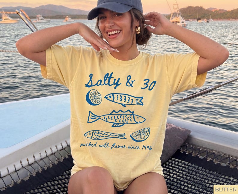 May include: A yellow t-shirt featuring the text "Salty & 30" and blue illustrations of fish and lemons. The shirt also has the text "packed with flavour since 1996".