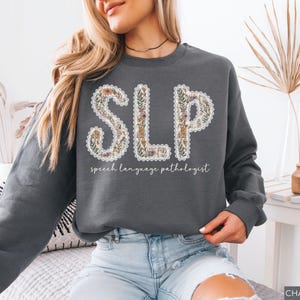 May include: Charcoal gray sweatshirt with the letters "SLP" in a floral design. The text "speech language pathologist" is below the letters. The sweatshirt is a long-sleeved crew neck.