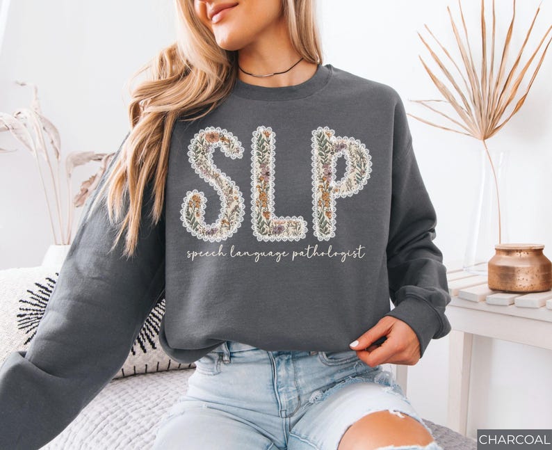 May include: Charcoal gray sweatshirt with the letters "SLP" in a floral design. The text "speech language pathologist" is below the letters. The sweatshirt is a long-sleeved crew neck.
