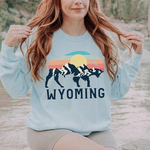 May include: A light blue sweatshirt with a graphic of a bison standing in front of a mountain range with a sunset behind it. The text "WYOMING" is printed below the graphic.