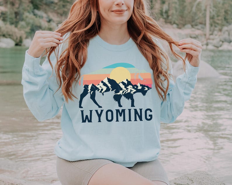 May include: A light blue sweatshirt with a graphic of a bison standing in front of a mountain range with a sunset behind it. The text "WYOMING" is printed below the graphic.