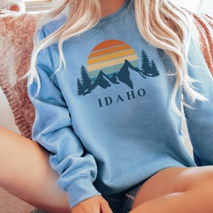 May include: A light blue sweatshirt with a graphic of a sunset over mountains and trees. The text "IDAHO" is printed below the graphic.