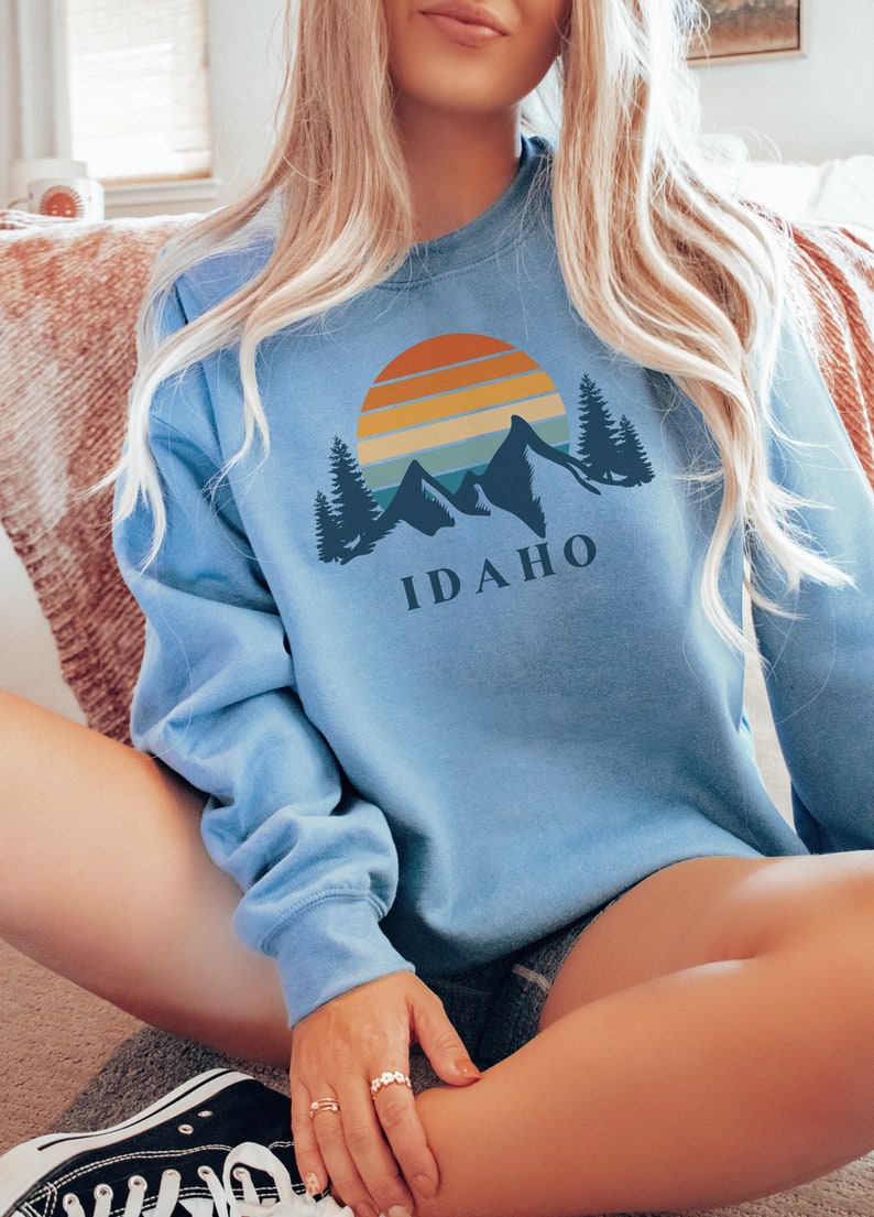 May include: A light blue sweatshirt with a graphic of a sunset over mountains and trees. The text "IDAHO" is printed below the graphic.