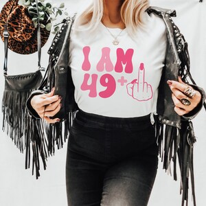 May include: White t-shirt with pink text that reads "I AM 49+" and a pink middle finger emoji.