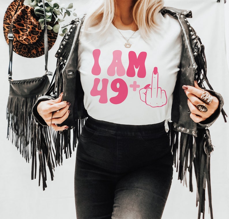 May include: White t-shirt with pink text that reads "I AM 49+" and a pink middle finger emoji.