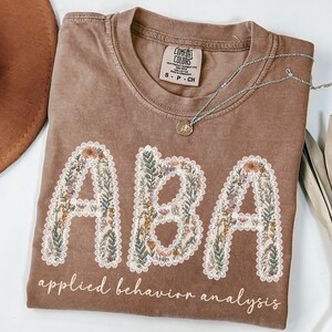 May include: A brown Comfort Colors t-shirt with the letters "ABA" in a floral design. The words "applied behavior analysis" are printed below the letters. A silver necklace with a pendant is on the shirt.