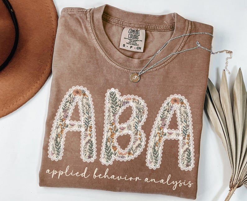 May include: A brown Comfort Colors t-shirt with the letters "ABA" in a floral design. The words "applied behavior analysis" are printed below the letters. A silver necklace with a pendant is on the shirt.