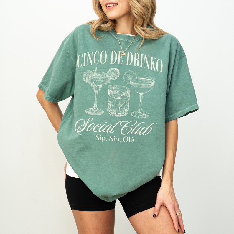 May include: A teal green t-shirt with a white graphic print of three cocktails and the text "Cinco De Drinko Social Club Sip, Sip, Ole".