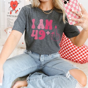 May include: A gray t-shirt with the text "I AM 49+" in pink letters. The number 49 is in a retro font and the plus sign is followed by a middle finger emoji.