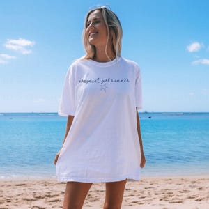 May include: A white oversized t-shirt with the text "pregnant girl summer" and a small starfish graphic printed on the front. The shirt is being worn by a person standing on a beach.