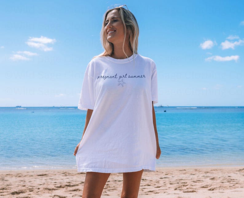May include: A white oversized t-shirt with the text "pregnant girl summer" and a small starfish graphic printed on the front. The shirt is being worn by a person standing on a beach.