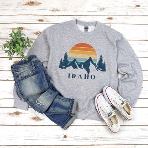 May include: A gray sweatshirt with a vintage-style graphic of mountains, a sunset, and the word "IDAHO". The sweatshirt is paired with blue jeans and white sneakers.
