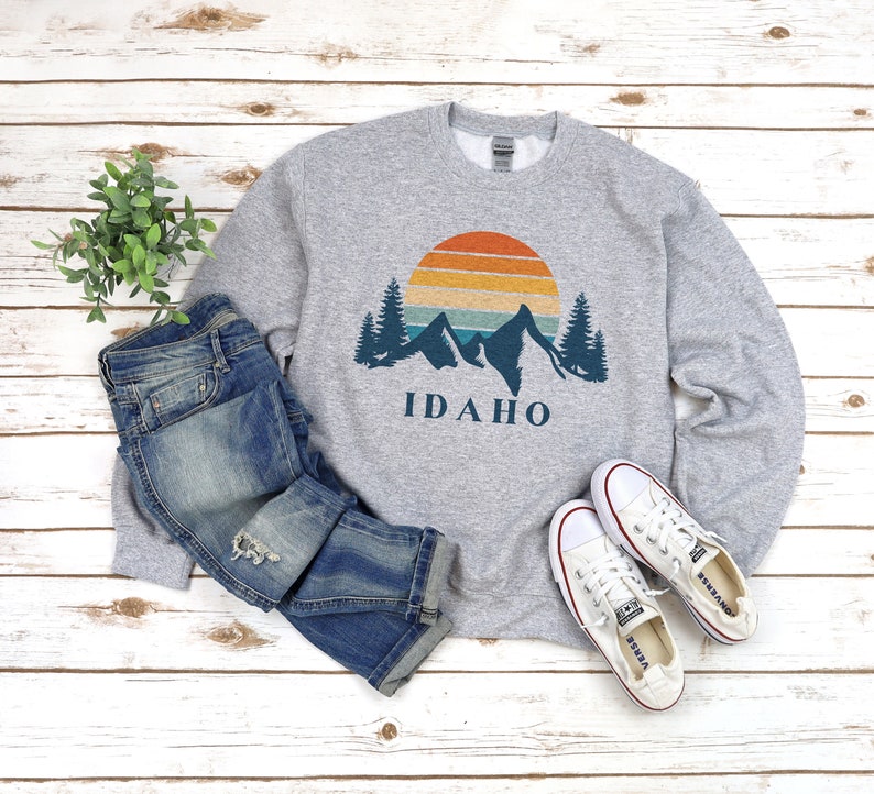 May include: A gray sweatshirt with a vintage-style graphic of mountains, a sunset, and the word "IDAHO". The sweatshirt is paired with blue jeans and white sneakers.
