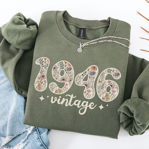 May include: An olive green sweatshirt with the year "1946" in a floral design and the word "vintage" below. A silver necklace with a small pendant is on the sweatshirt. The sweatshirt is folded on top of a pair of blue jeans.