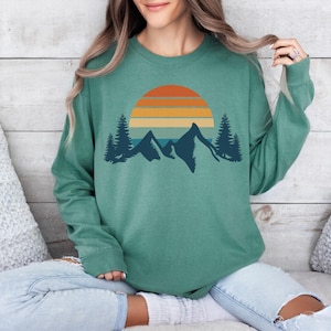 May include: A teal green long-sleeved sweatshirt with a retro sunset and mountain graphic. The graphic features a multi-colored sunset with orange, yellow, and blue stripes, and a silhouette of mountains with trees in front.