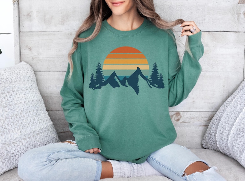May include: A teal green long-sleeved sweatshirt with a retro sunset and mountain graphic. The graphic features a multi-colored sunset with orange, yellow, and blue stripes, and a silhouette of mountains with trees in front.