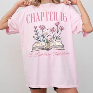 Puede incluir: Camiseta rosa de gran tama&ntilde;o con un gr&aacute;fico de un libro abierto con flores que salen de &eacute;l. El texto "CHAPTER 16" est&aacute; por encima del libro y "A Literary Milestone" est&aacute; por debajo del libro.