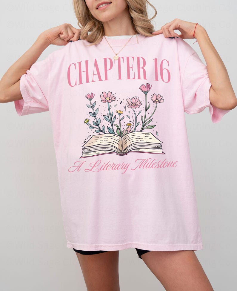 Puede incluir: Camiseta rosa de gran tama&ntilde;o con un gr&aacute;fico de un libro abierto con flores que salen de &eacute;l. El texto "CHAPTER 16" est&aacute; por encima del libro y "A Literary Milestone" est&aacute; por debajo del libro.