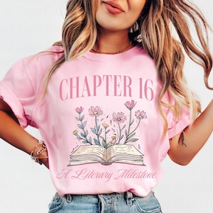 Puede incluir: Camiseta rosa con el texto "Cap&iacute;tulo 16 Un Hito Literario" y un dibujo de un libro abierto con flores.