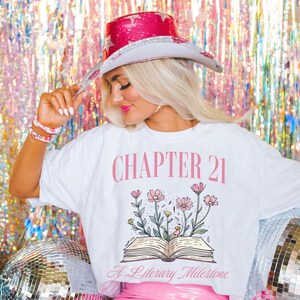 May include: White t-shirt with a pink graphic of an open book with flowers growing out of it. The text "CHAPTER 21" is above the book and "A Literary Milestone" is below the book.