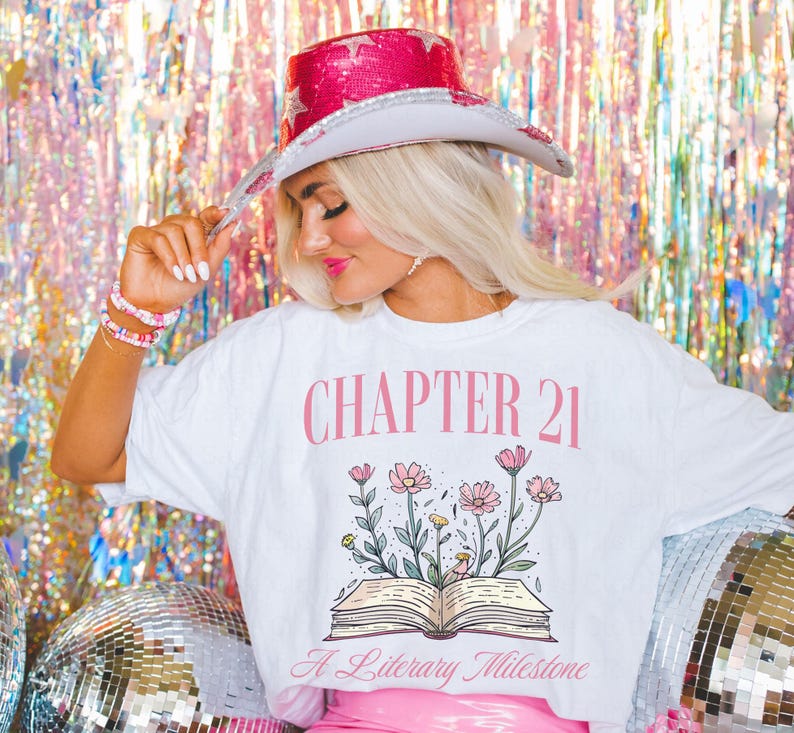 May include: White t-shirt with a pink graphic of an open book with flowers growing out of it. The text "CHAPTER 21" is above the book and "A Literary Milestone" is below the book.