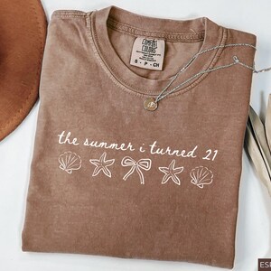 Pu&ograve; includere: T-shirt color espresso con la scritta "the summer i turned 21" e illustrazioni di conchiglie, stelle marine e fiocchi. Visibile anche una collana d'argento con un ciondolo e un cappello marrone.