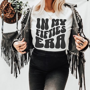 May include: White t-shirt with black text that reads "In My Fifties Era" in a retro font. The person is wearing a black leather jacket with fringe.