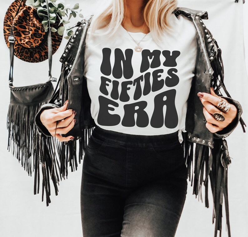May include: White t-shirt with black text that reads "In My Fifties Era" in a retro font. The person is wearing a black leather jacket with fringe.